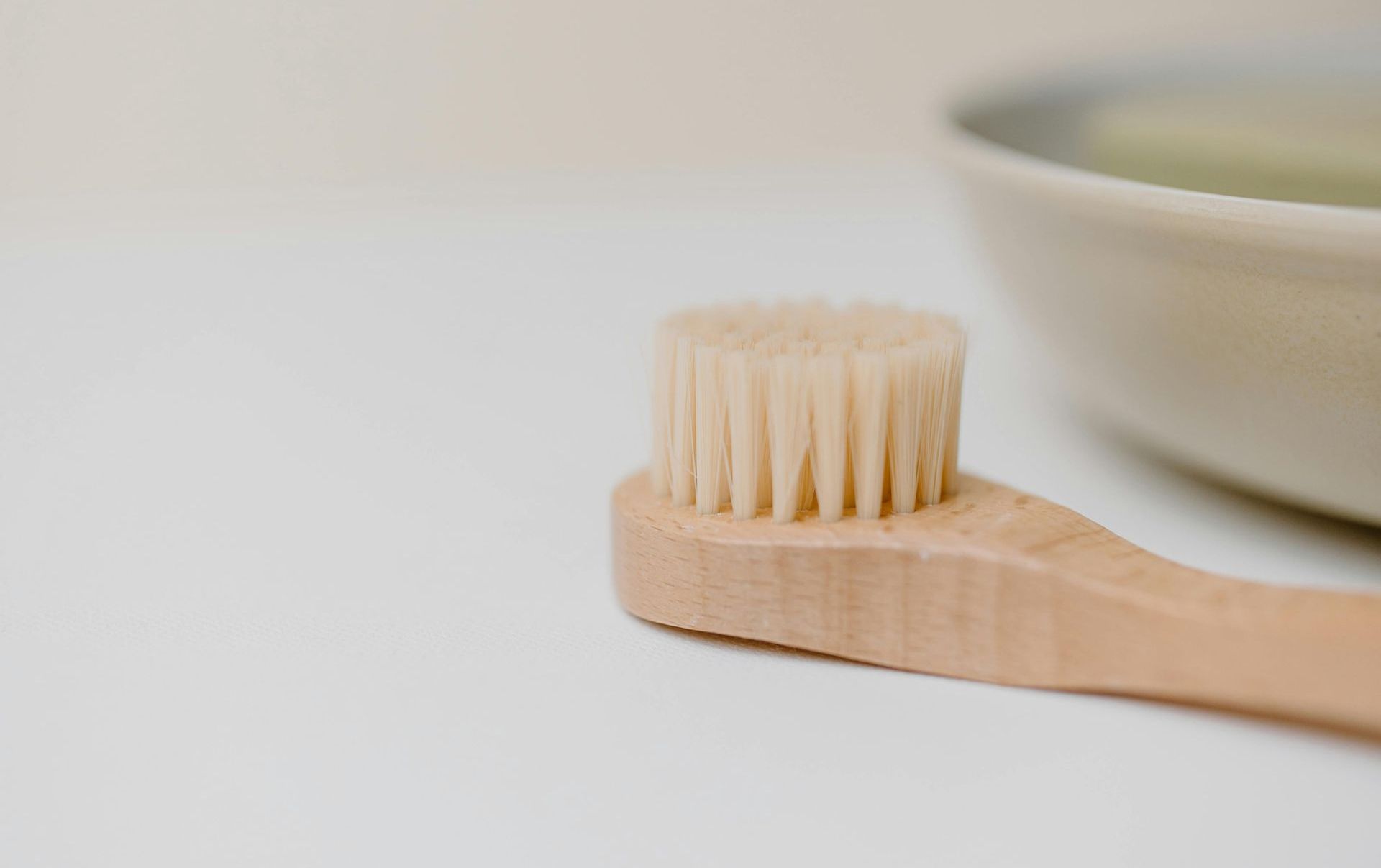 A wooden exfoliating brush is sitting on a white table next to a bowl of soap.