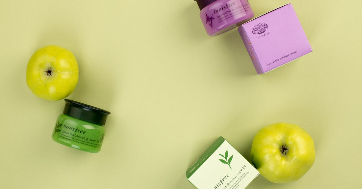 A yellow apple , a green apple , a purple box , and a green cream are on a table.