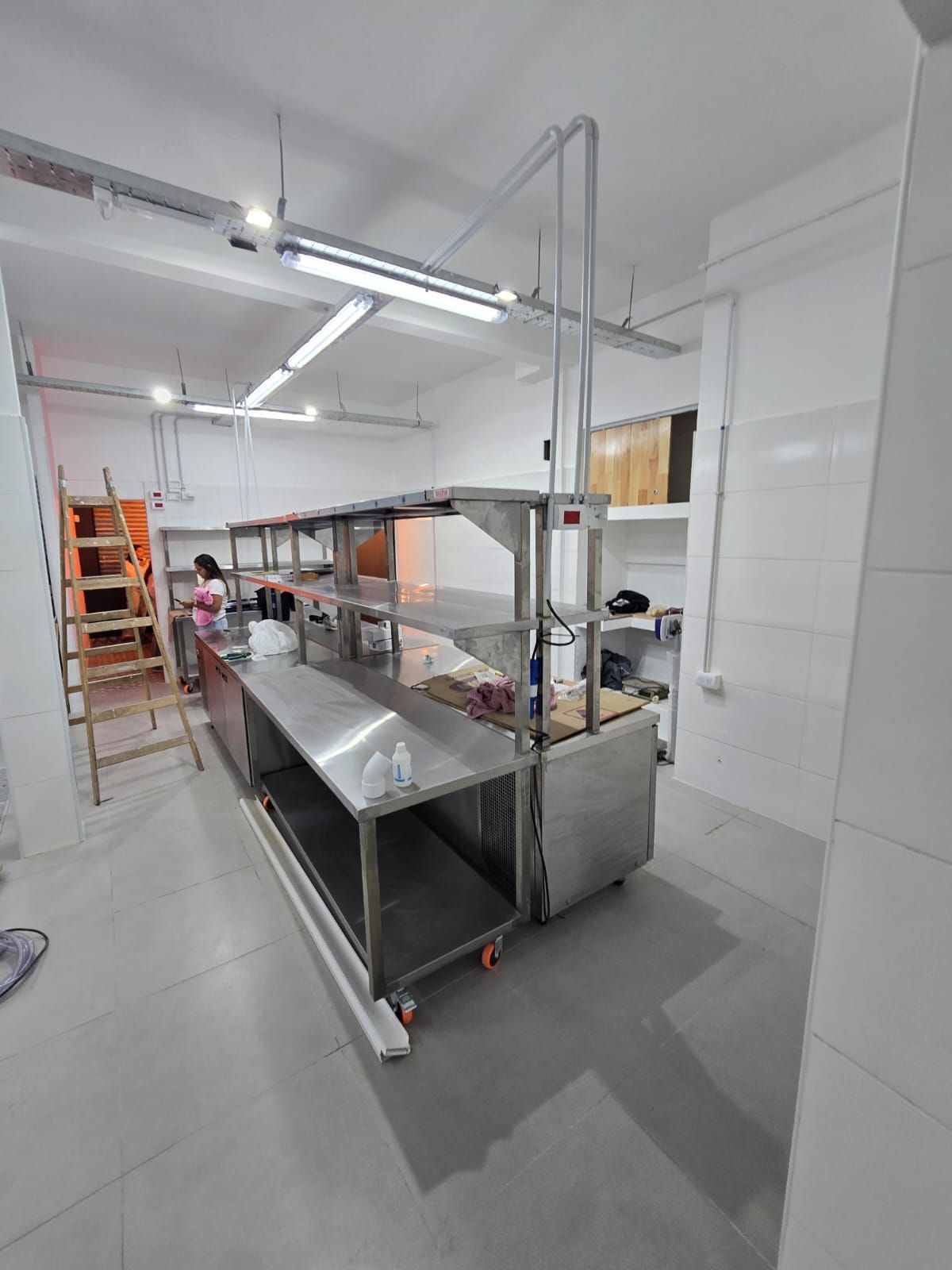 Cocina industrial luminosa con mesas de preparación de acero inoxidable, estantes y una escalera bajo