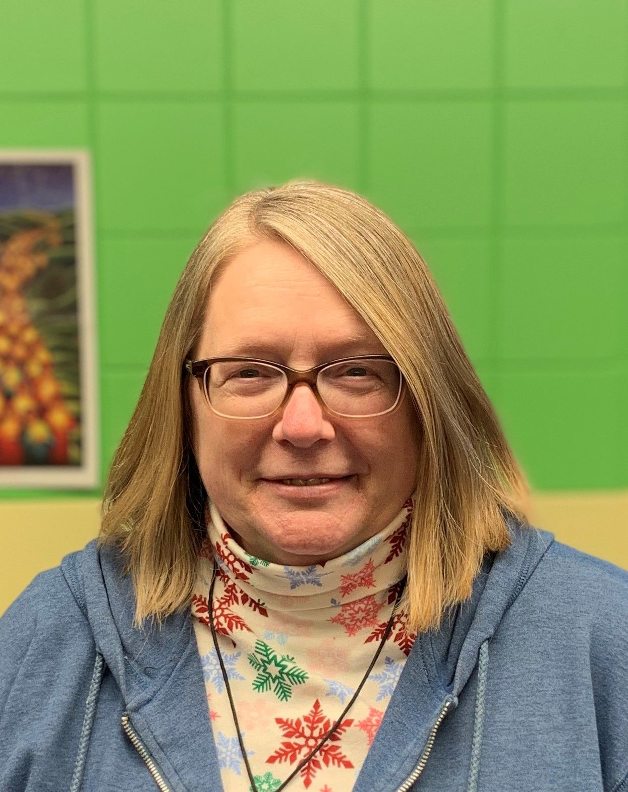 A woman wearing glasses and a blue hoodie is smiling in front of a green wall.