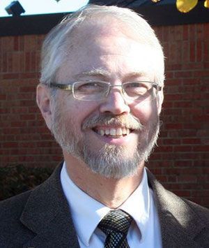 A man with glasses and a beard is smiling in front of a brick wall