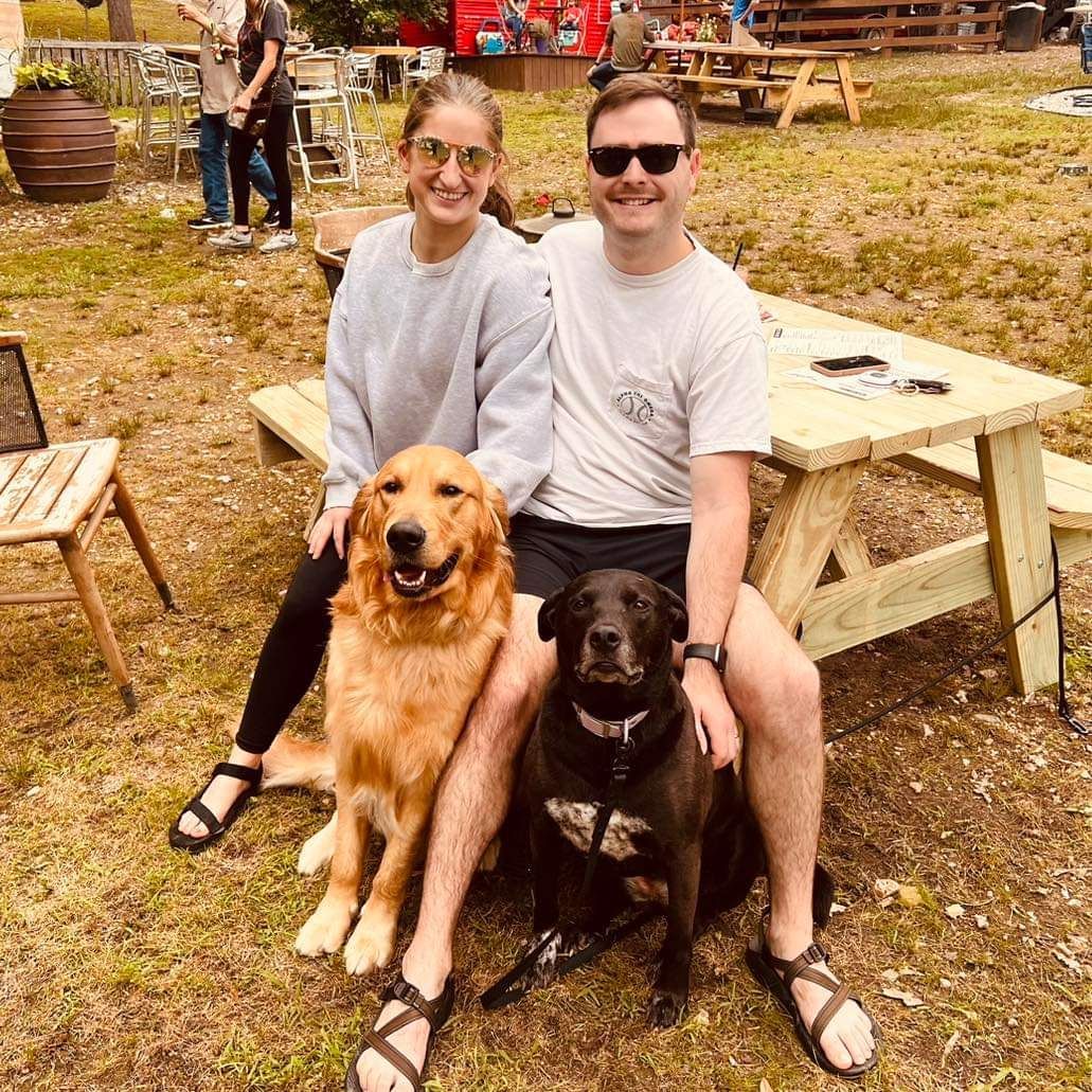 A man and a woman are sitting on a picnic table with two dogs.