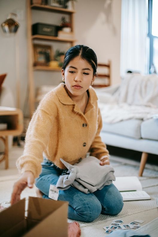 Woman in yellow sweater and jeans packing clothes into a cardboard box on the floor.