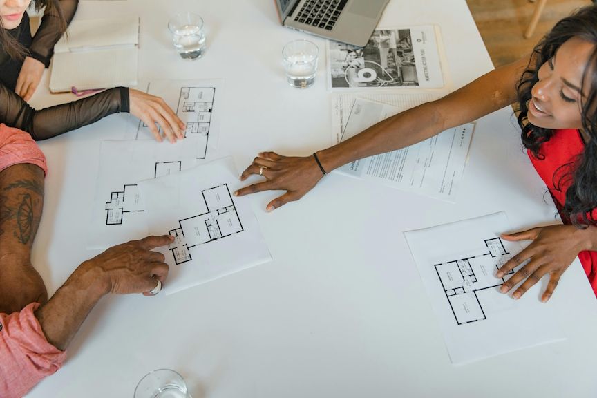 People reviewing architectural floor plans at a white table. One person in red, others in pink and black. Laptop present.