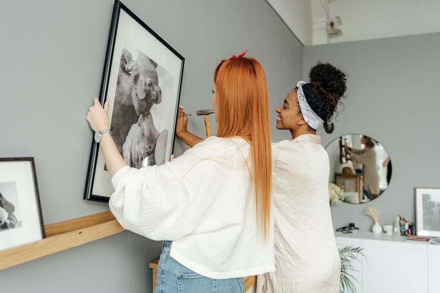 Two people hanging a framed picture on a gray wall. One uses a hammer.