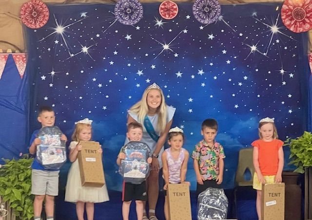 A woman wearing a crown poses with six children, each holding a box or bag, in front of a blue starry backdrop.