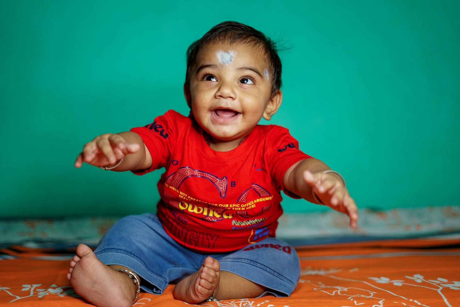 A smiling child in a red shirt and blue jeans sitting on an orange surface against a bright turquoise background.