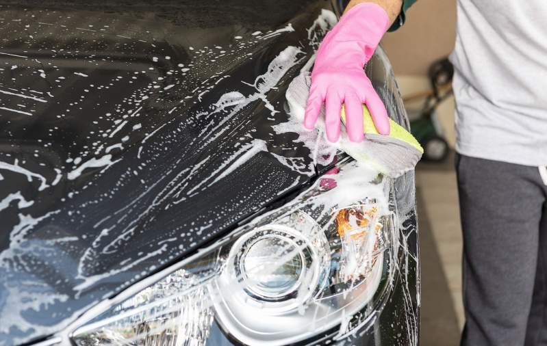 Una persona lava un'auto nera con la schiuma di sapone; una mano con un guanto rosa pulisce il cofano e il faro.