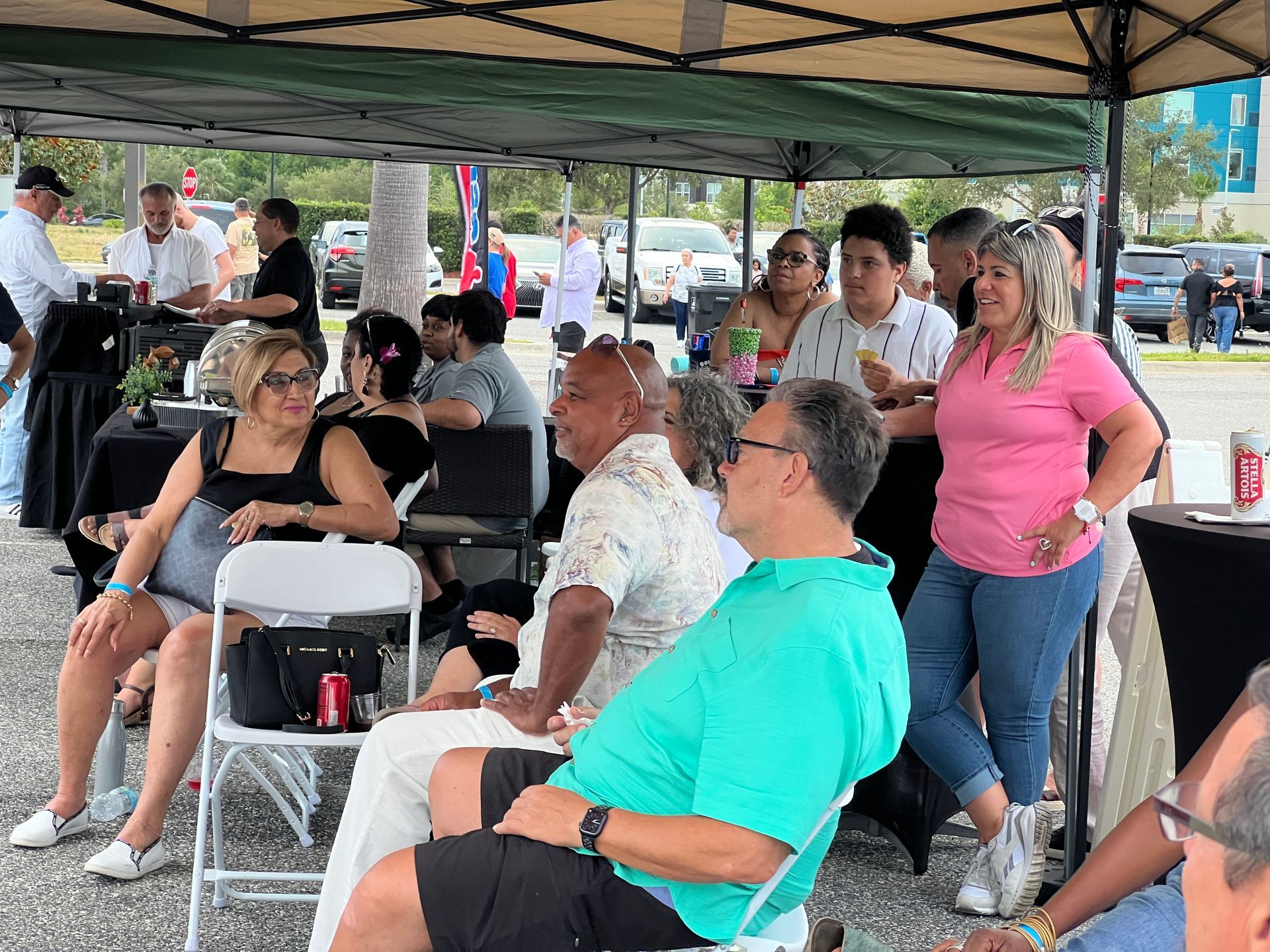 People gathered outdoors under a tent, some seated, socializing and drinking. Cars in the background.