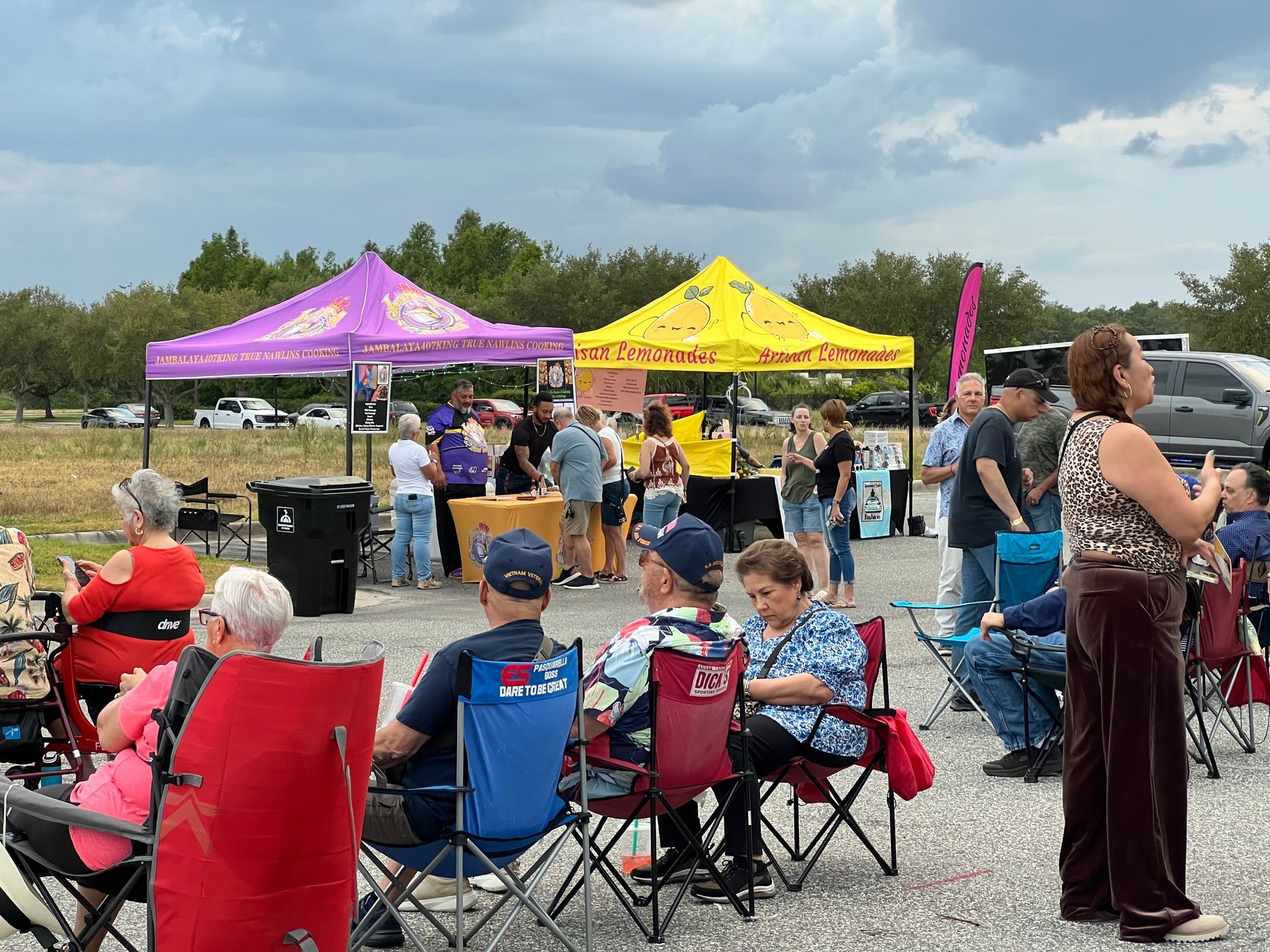Outdoor market with vendor tents, people browsing, and people seated in folding chairs.