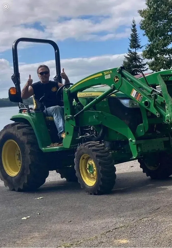 Man giving thumbs up, seated on a green John Deere tractor with a loader, near water.