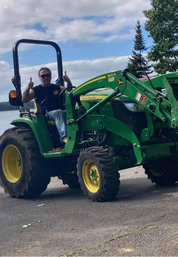 Man on a green John Deere tractor gives thumbs up, outdoors by water.