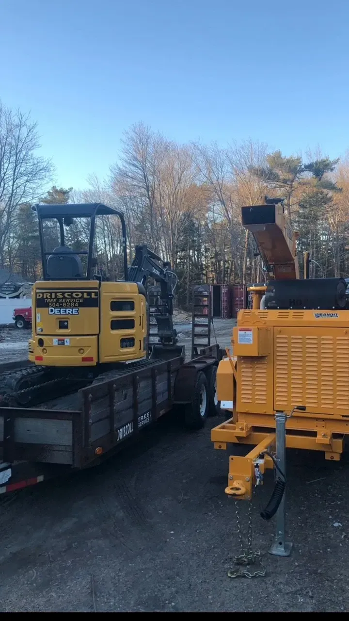 Yellow excavator on a flatbed trailer next to a wood chipper, outdoors.