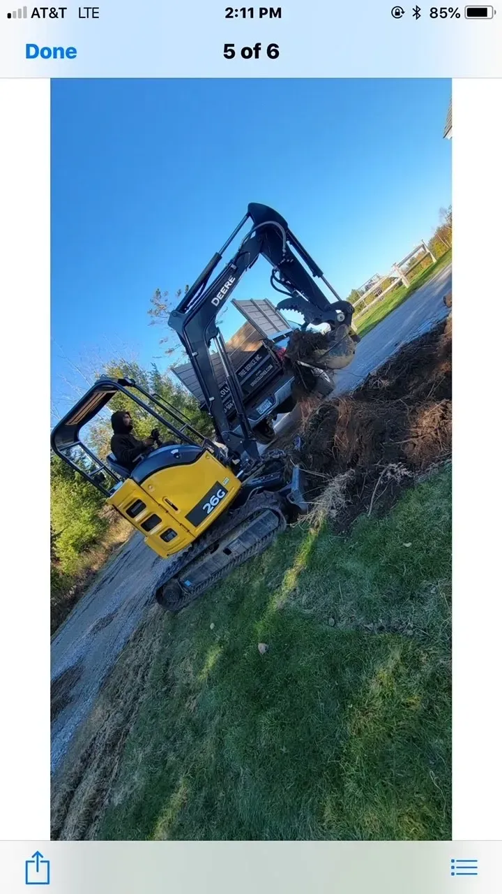 Yellow excavator digging on a grassy slope next to a road under a blue sky.