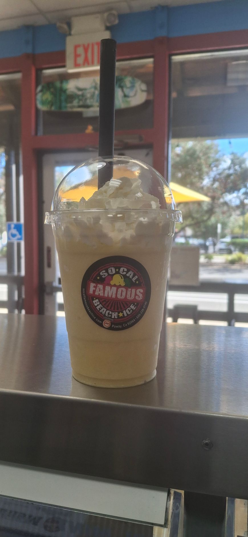 A milkshake in a plastic cup with whipped cream and a straw on a counter near a window.