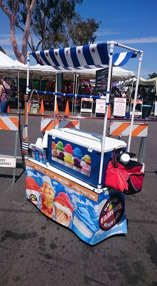 Ice cream cart with blue and white striped awning and ice cream cone graphics, parked on pavement.