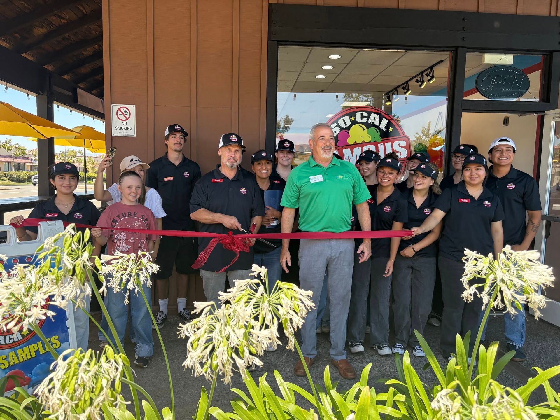 People cutting ribbon at the grand opening of a So Cal restaurant, with staff, flowers, and building entrance.