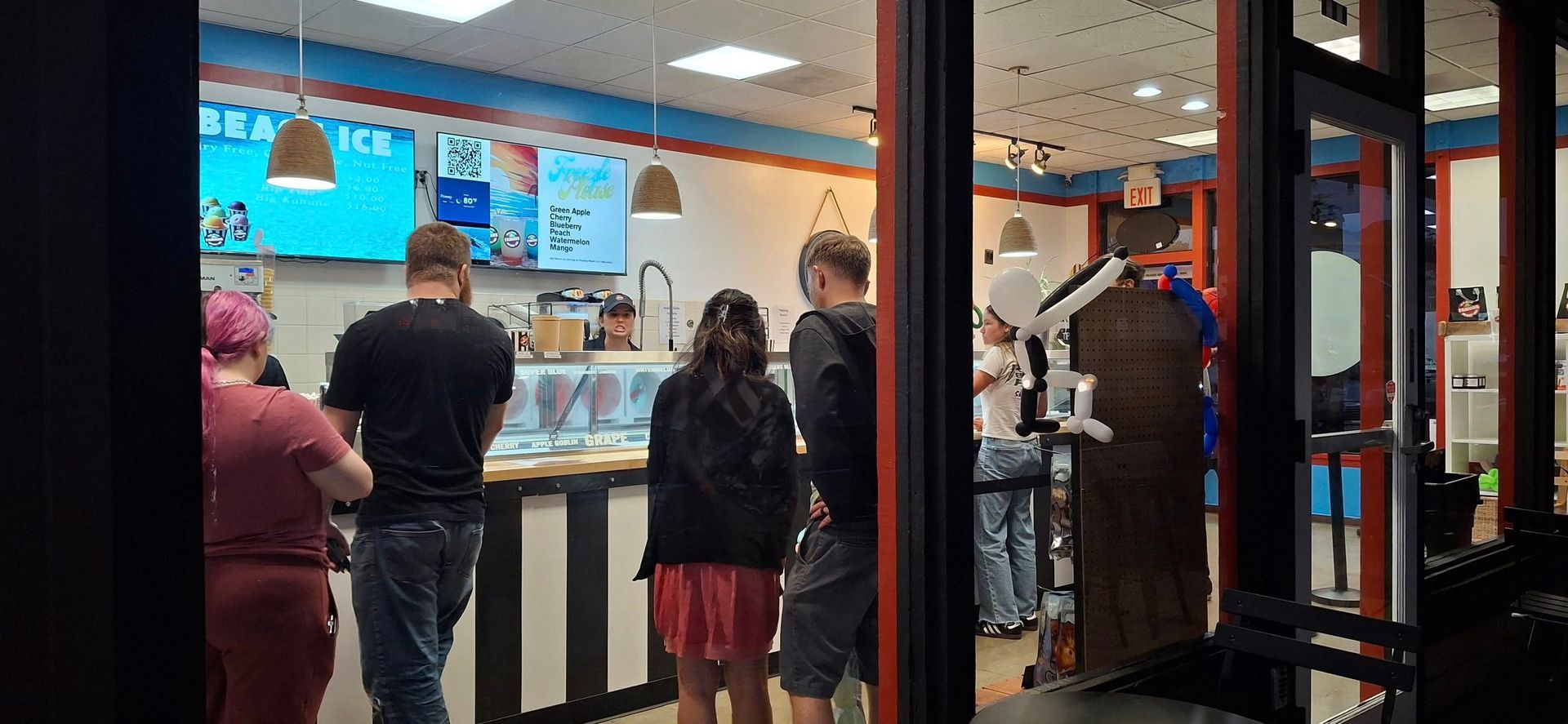 People inside a brightly lit ice cream shop. Customers stand in line at the counter. A few people stand inside the doorway.
