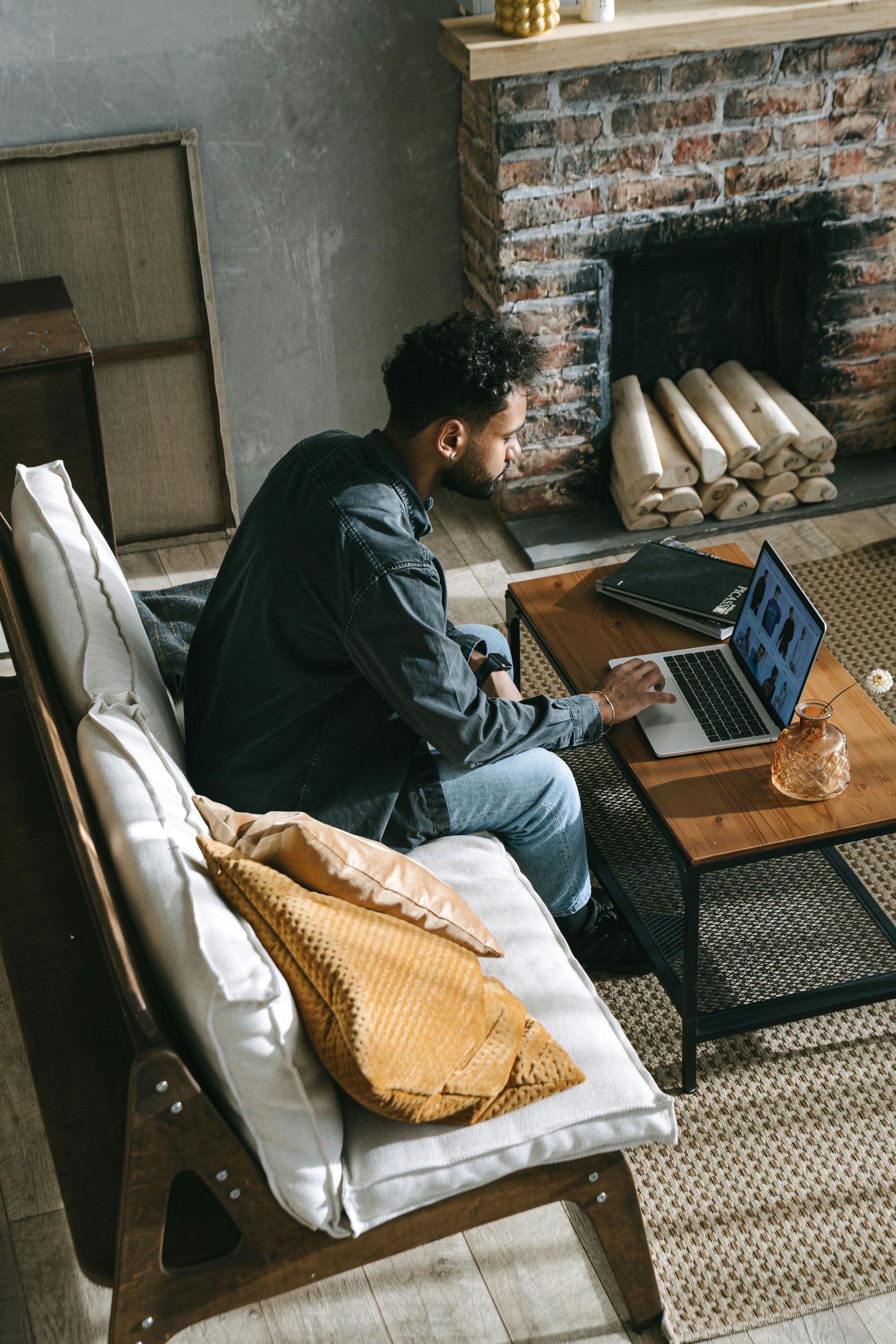 Man working on a laptop on a coffee table near a couch by a fireplace.