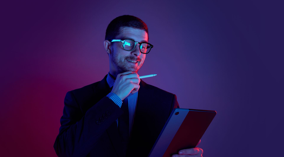 Man in a suit, holding a tablet and pen, looking thoughtful under red and blue lights.