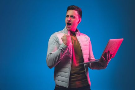 Man clenching fist, celebrating with open mouth, holding laptop. Pink and blue neon lighting on blue background.