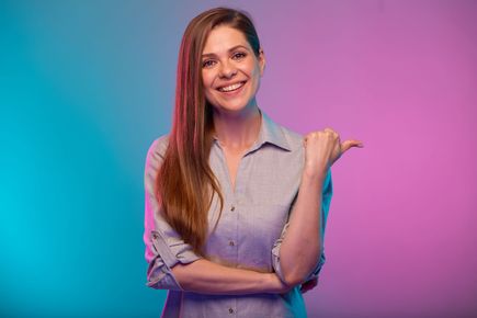 Woman with long brown hair smiles and points right, against blue and pink backdrop.
