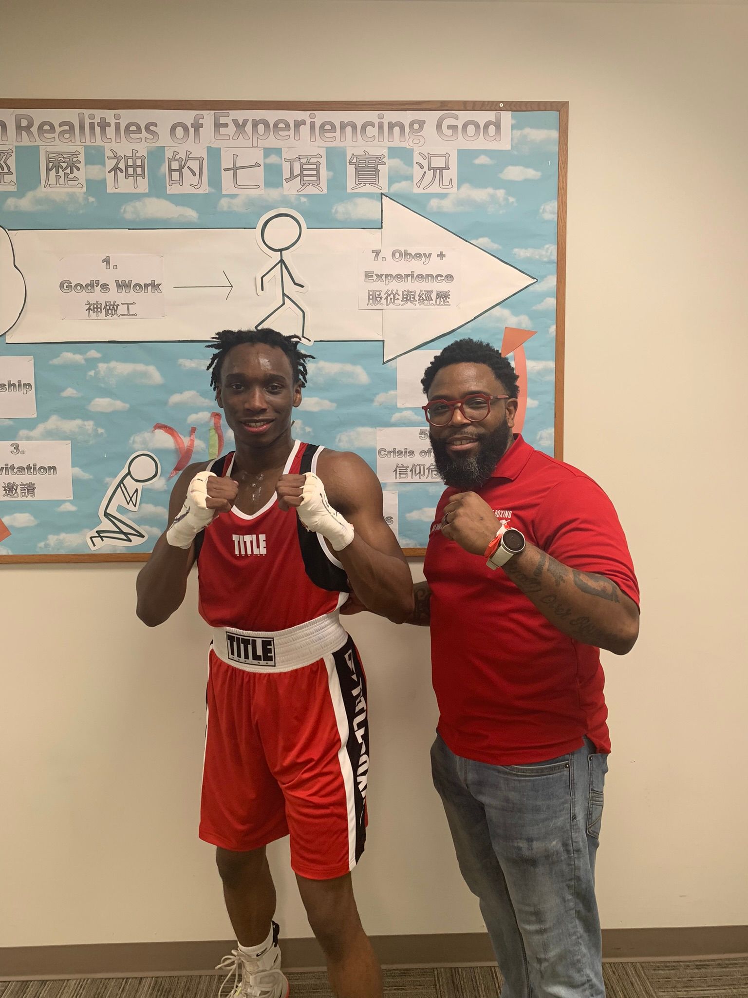 Two men are standing next to each other in front of a bulletin board.