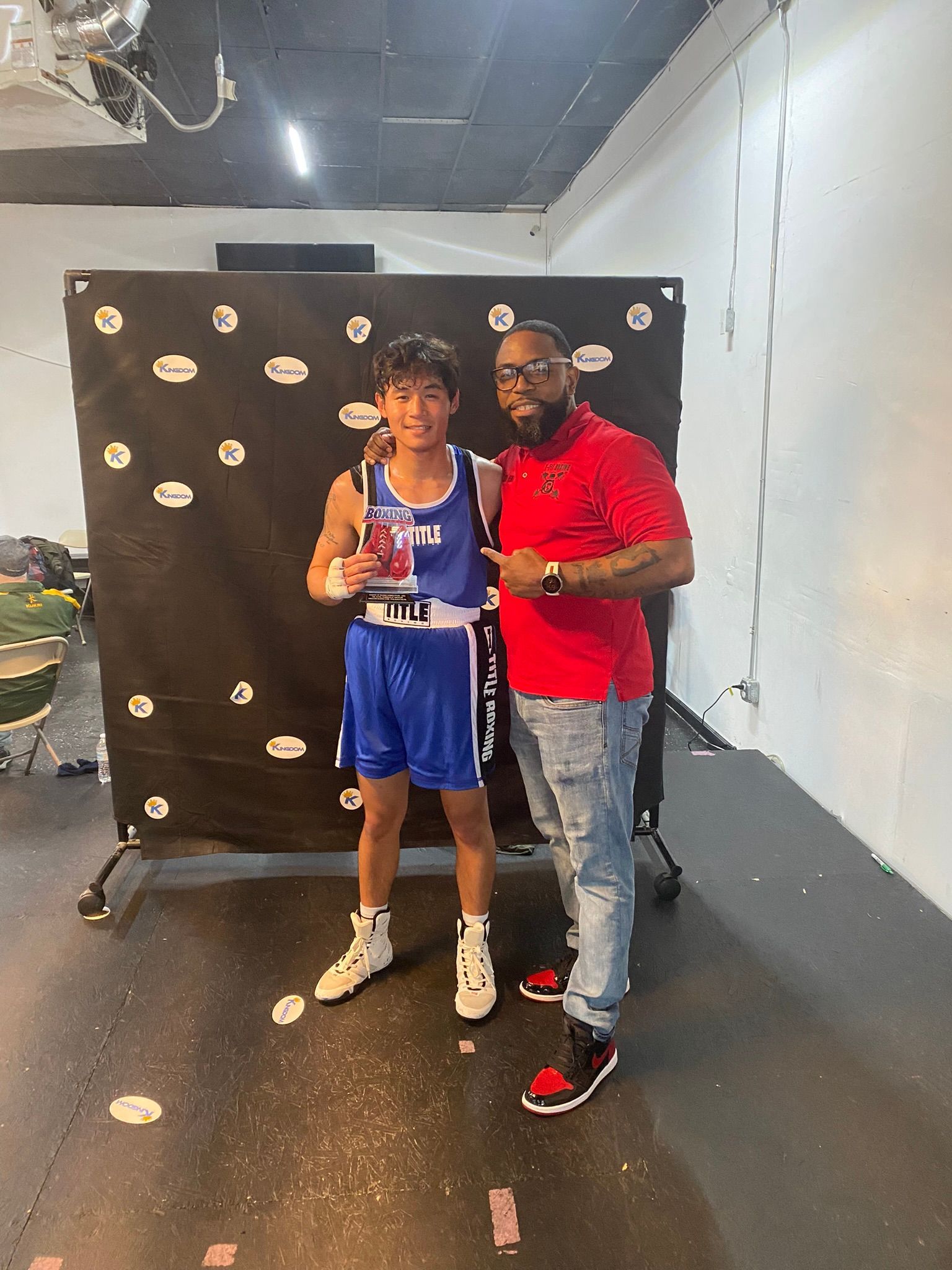 A man in a red shirt is standing next to a young boy in a blue boxing uniform.