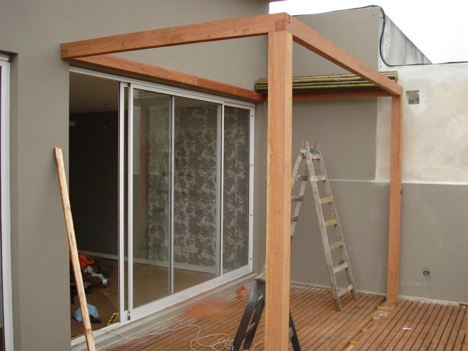 Pérgola de madera en construcción junto a puertas corredizas de vidrio en una terraza. Una escalera se apoya en la pared.