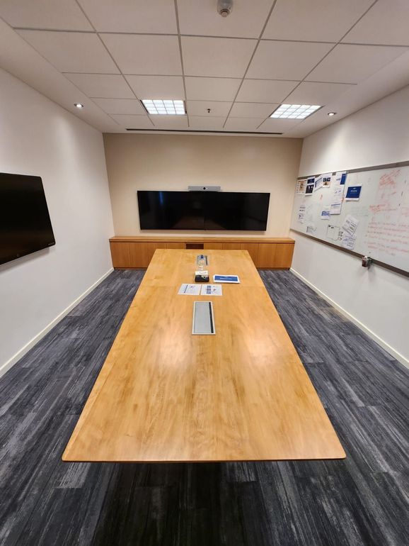 Sala de conferencias con mesa larga de madera, pantalla grande, pizarra y alfombra azul.