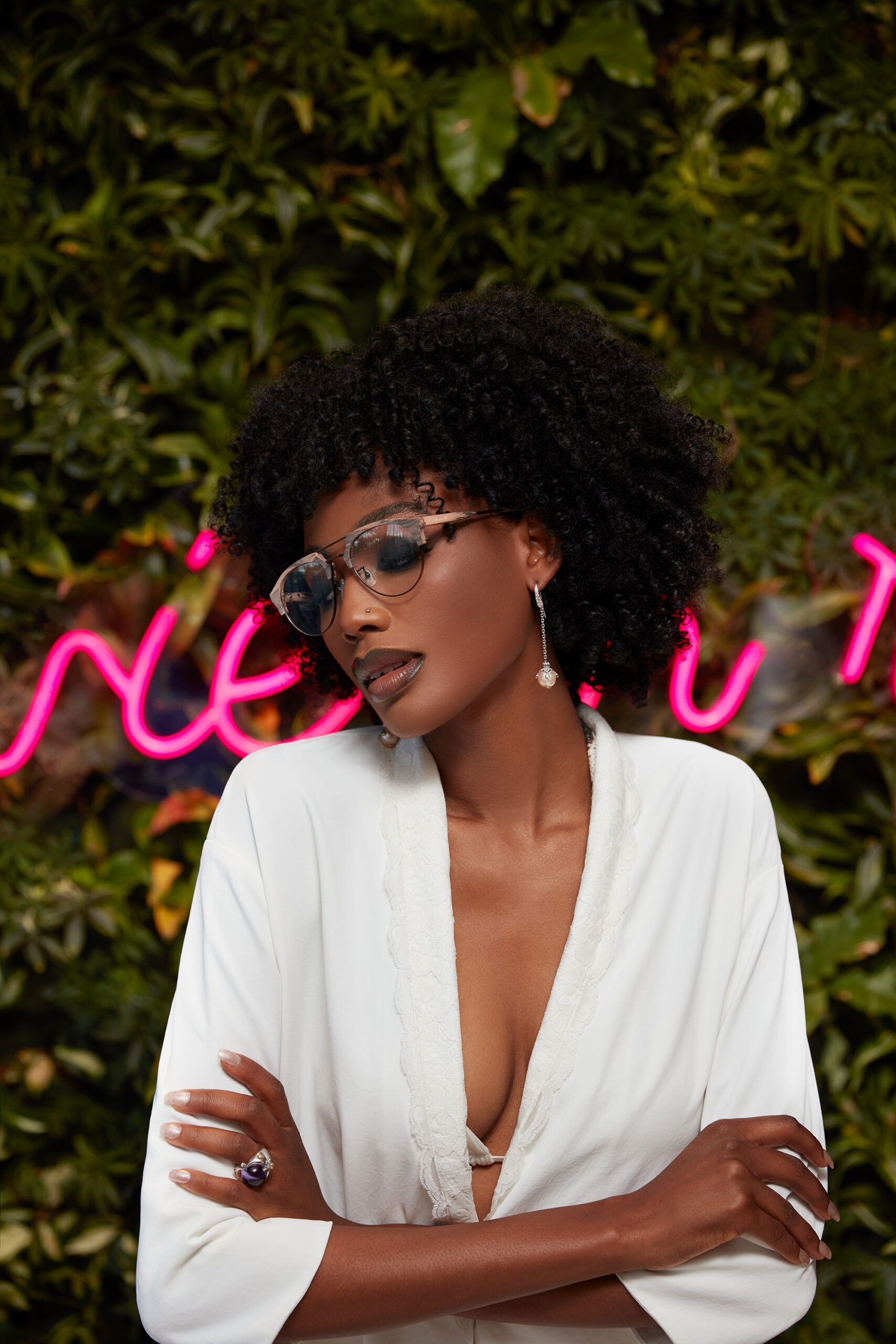 A woman wearing sunglasses and a white shirt is standing in front of a neon sign.