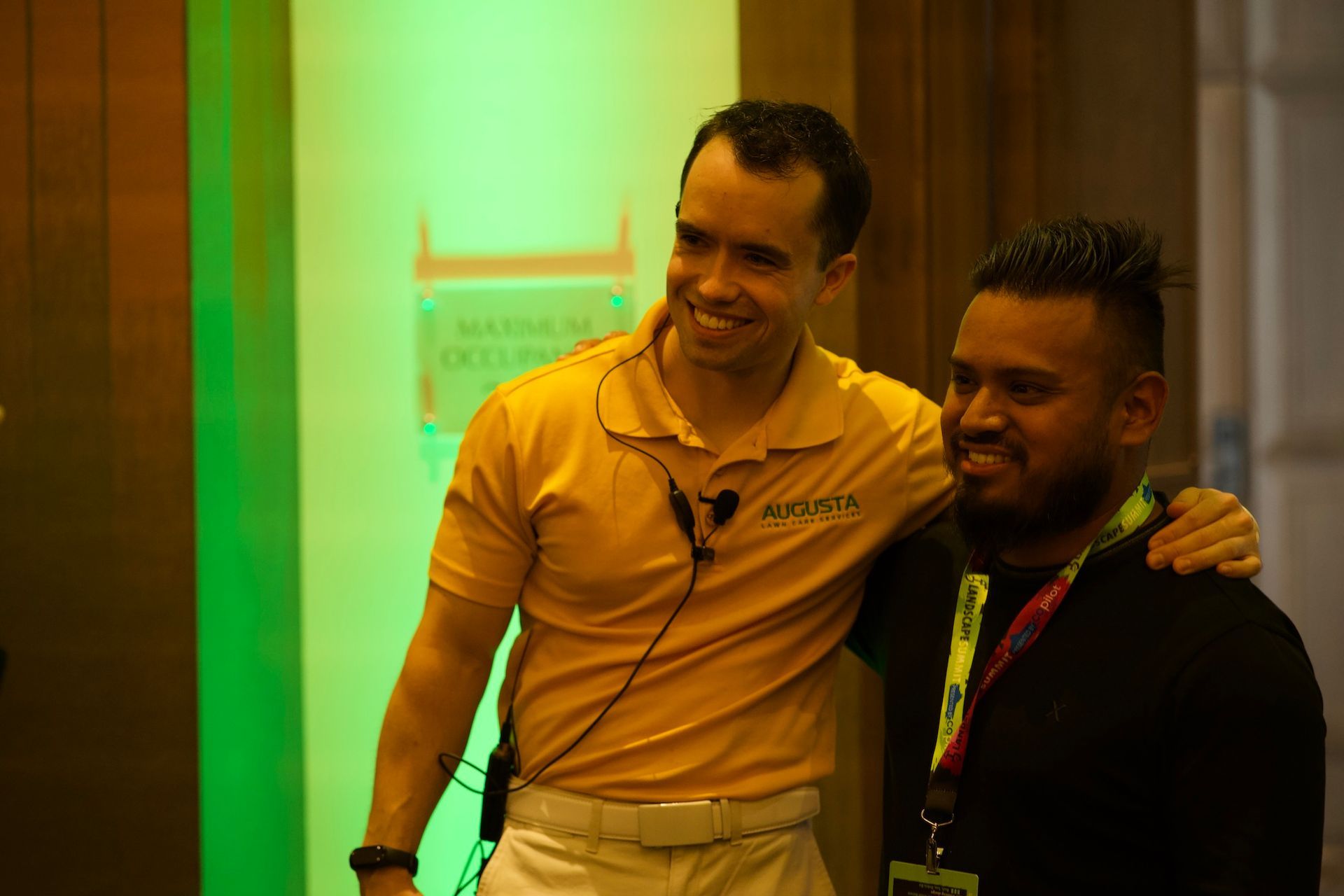 Two men are posing for a picture in front of a green wall.