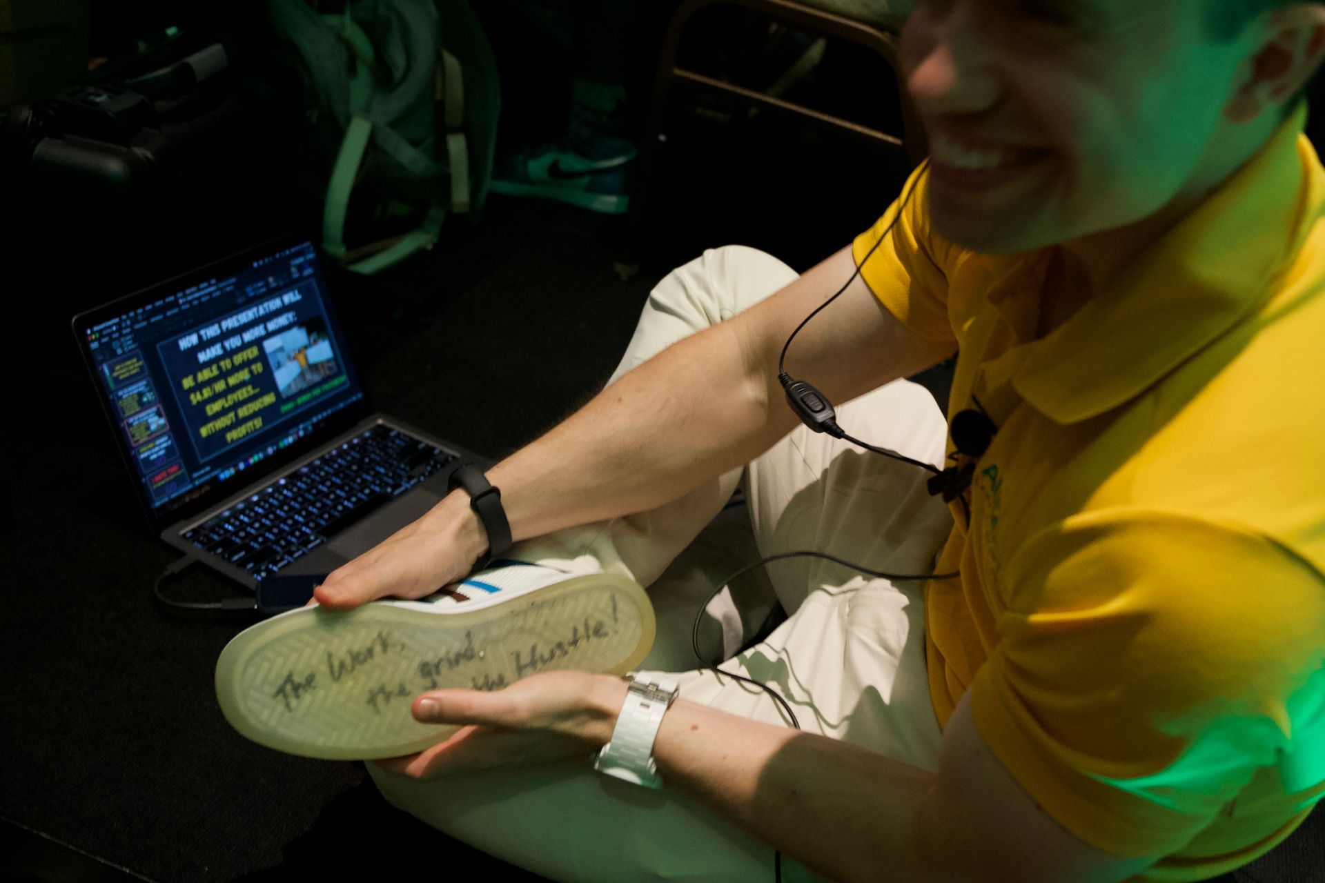 A man in a yellow shirt is holding a shoe in front of a laptop.