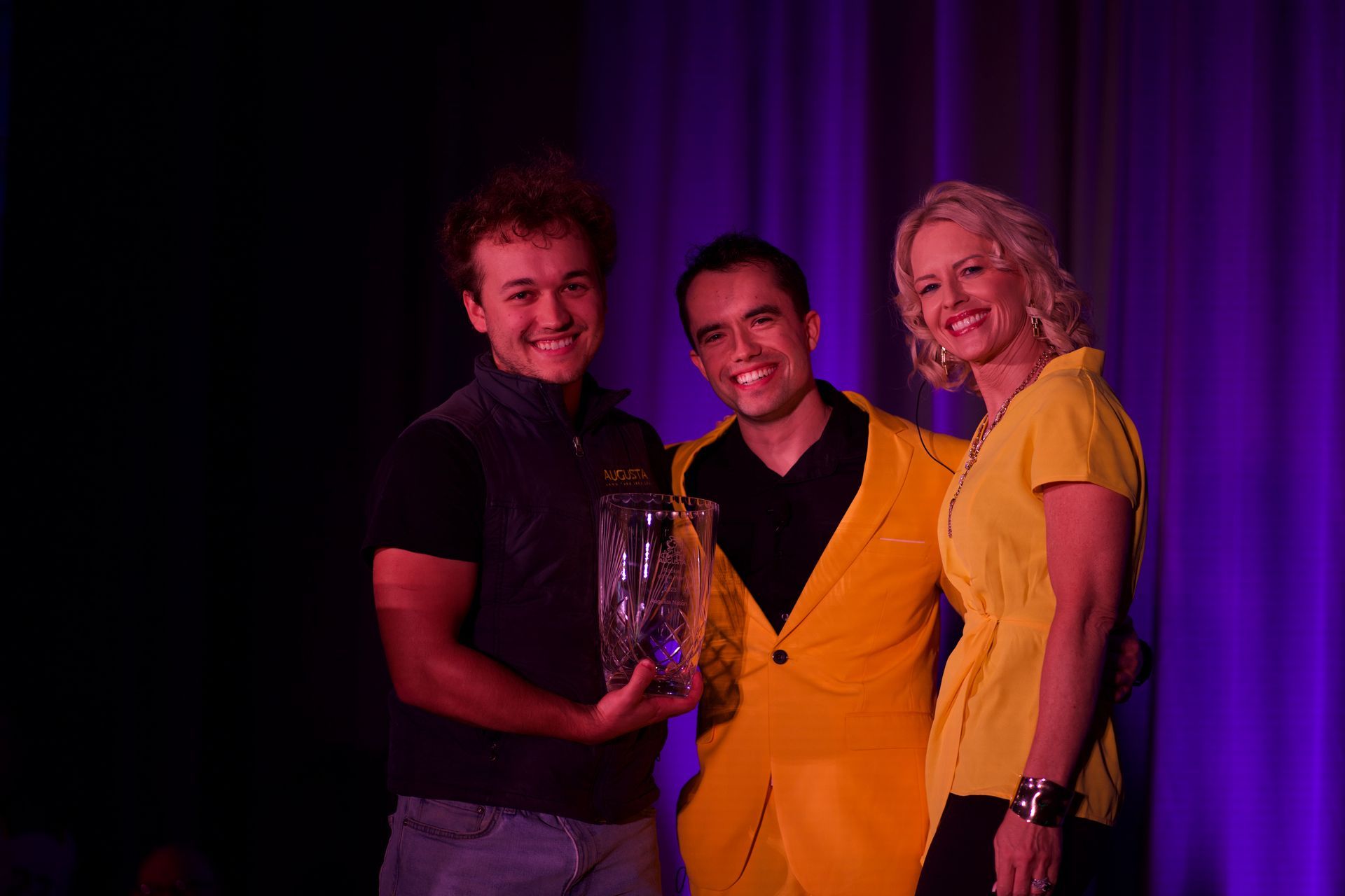 A man in a yellow suit is holding a trophy while standing next to two women.