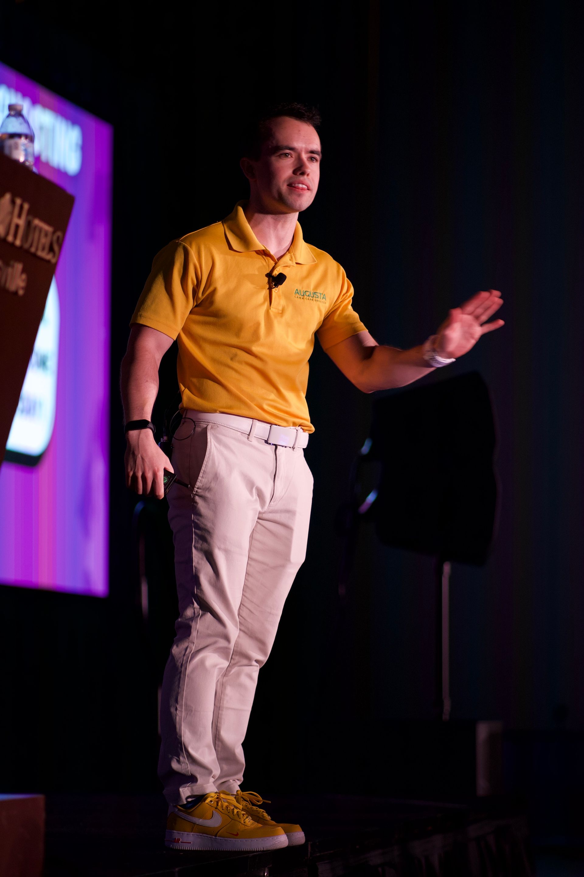 Mike Andes in a yellow shirt is standing on a stage giving a presentation.