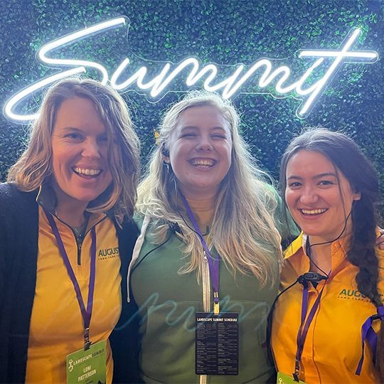 Three women are posing for a picture in front of a neon sign that says summit