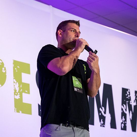 Tommy Mello is in a black shirt holding a microphone in front of a sign that says summit, talking to a large group