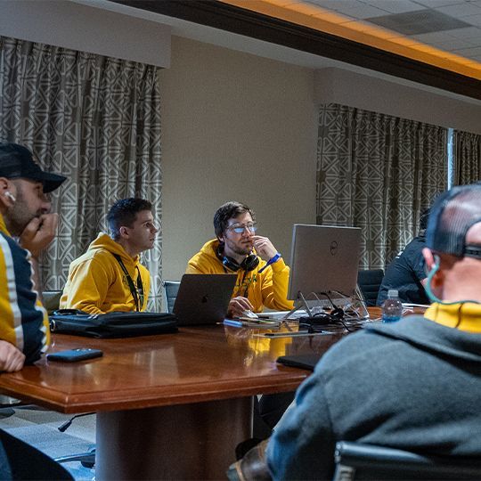 A group of men are sitting around a table with laptops.