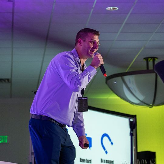 Tommy Mello is talking into a microphone while standing on a stage at summit