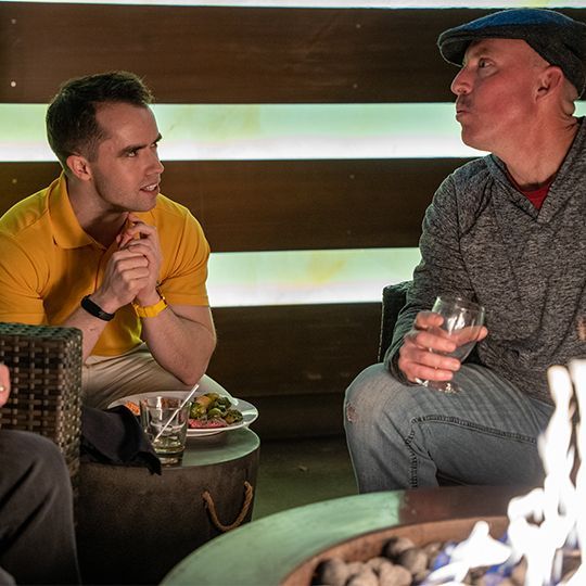 Two men are sitting around a fire pit talking to each other