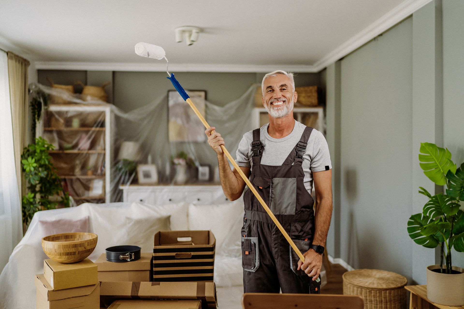 A painter holds a painting roller and prepares for painting residential walls. Covered furniture is behind him. A painter holds a painting roller and prepares for painting residential walls. Covered furniture is behind him.