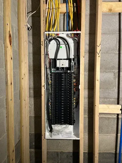 Electrical panel installed in a wooden frame against a gray cinder block wall; wires and breakers are visible.