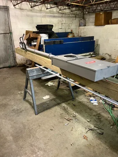 A cluttered basement with a sawhorse, electrical panel, and various materials.