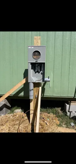A gray electrical meter box mounted on wooden supports against a green wall; dry grass and dirt in the foreground.