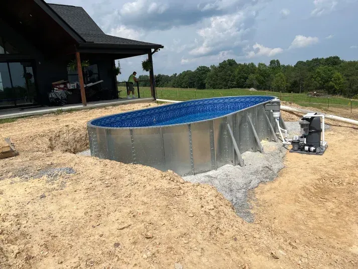 Oval above-ground pool under construction next to a house. Partially dug into the ground.