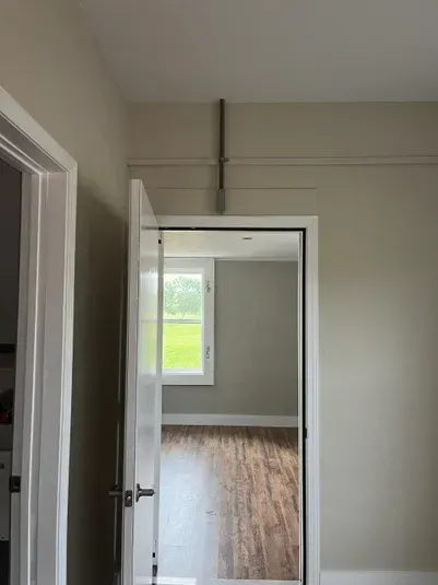 A doorway with exposed pipes running across the top. Beyond the doorway is a room with a window and wood flooring.