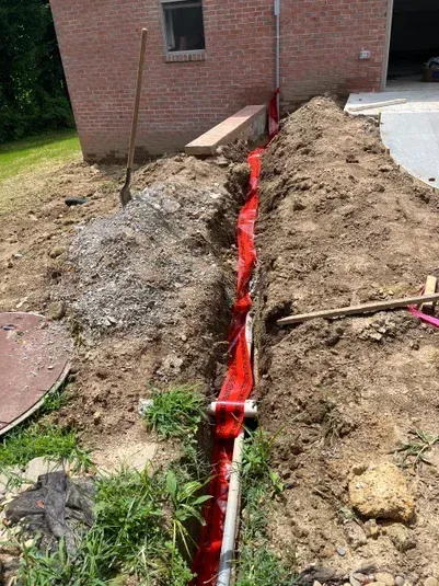 Trench along a brick building with a red liner, gravel, and a shovel.