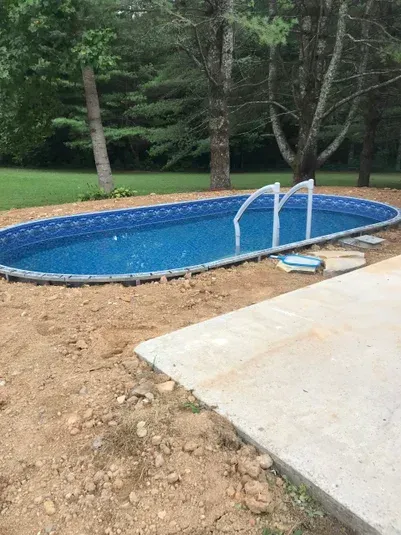 Oval swimming pool with a blue interior, surrounded by dirt, concrete, and trees.