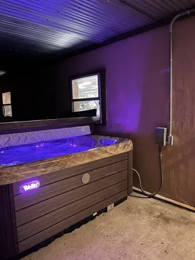 A hot tub with blue lights under a corrugated metal roof, near a window and wall with a power outlet.