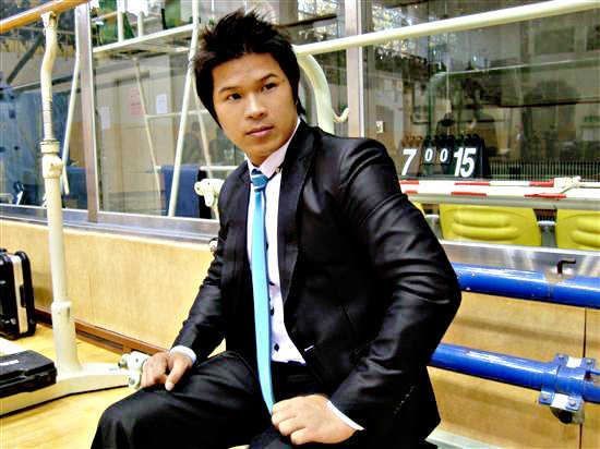 Man in a black suit with a blue tie, seated, posing in front of a gym setting.